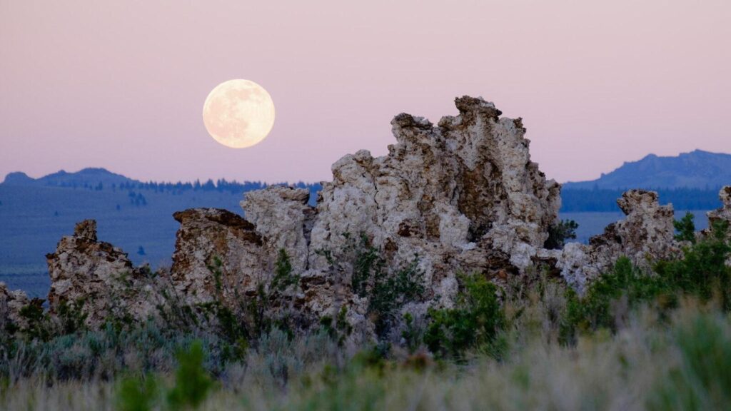 A photograph of a pink sky, full moon, and a rocky landscape.