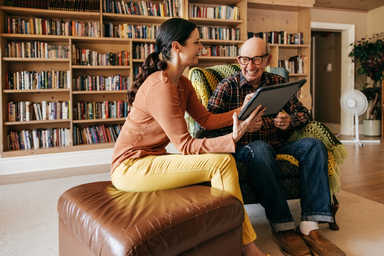 A caregiver and an elderly man are sitting on a couch in a library. The caregiver is gesturing on a large tablet to show the senior man something.