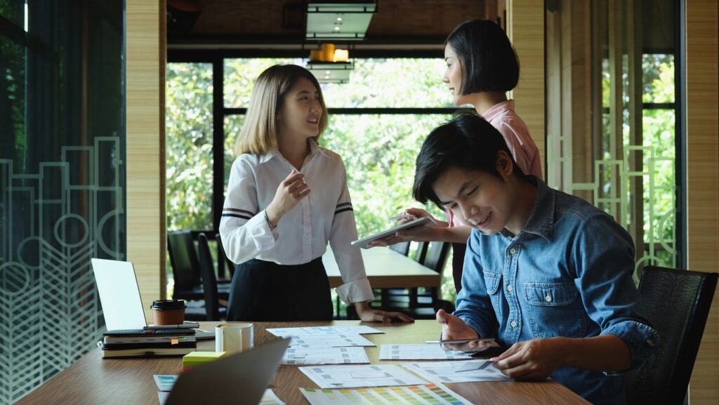 A group of young adults having a discussion in an office, taking notes on paper and viewing a laptop at the edge of the desk.