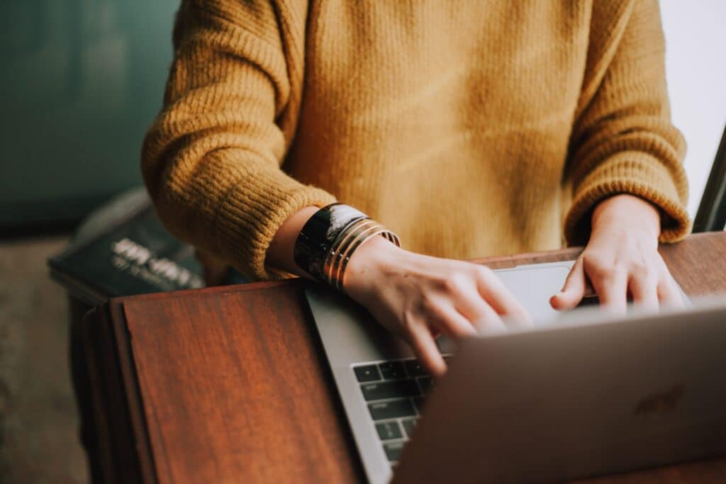 woman in yellow sweater sitting and working on laptop on a wooden desk.