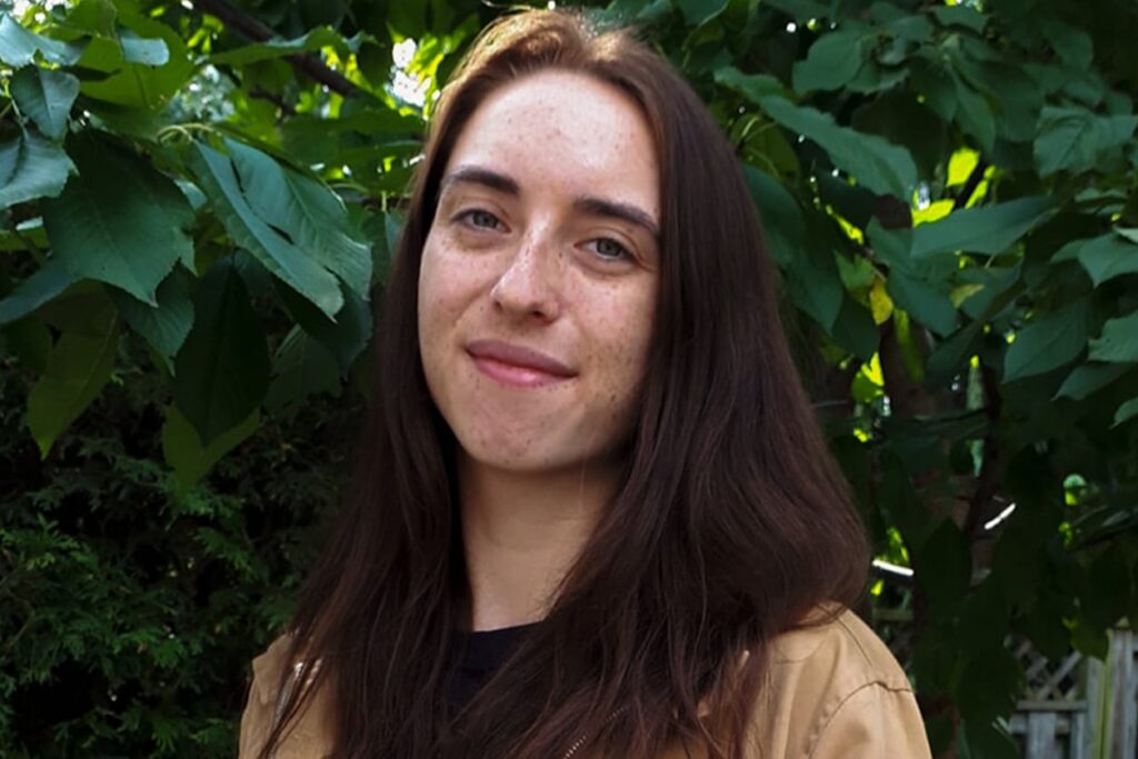 Young woman with brown hair photographed outdoors in front of foliage.